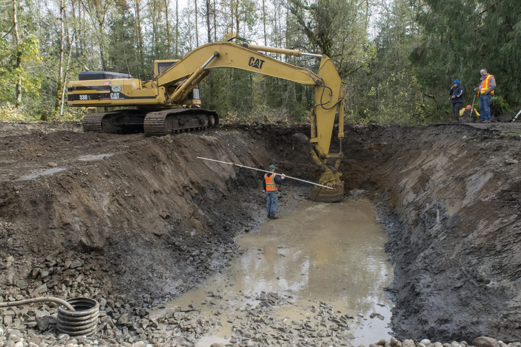 Photo of an excavator digging up a hole filled with water. There are two other workers looking down in the hole while another is holding a long rod in the hole.