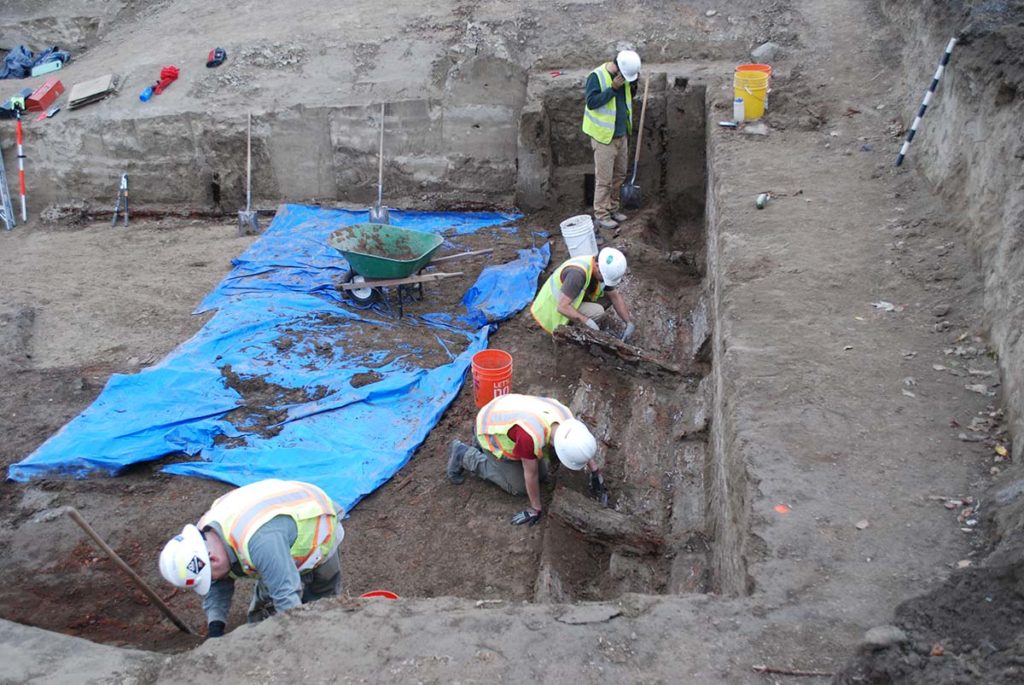 Photo of workers digging in a site. There is a large blue tarp where workers are putting what they dig up on.