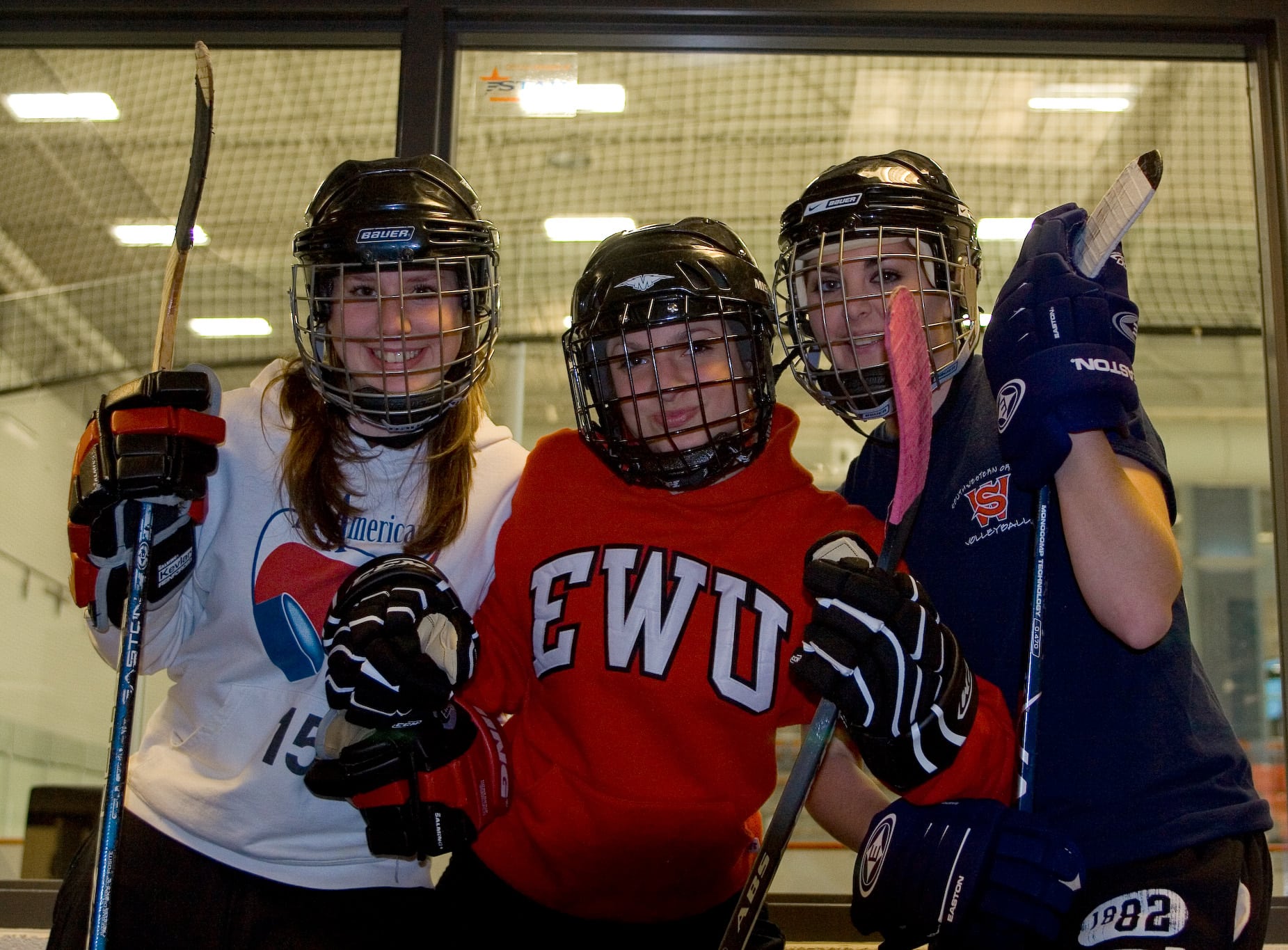 Baitlyn, Kayla, and Daitlin pose, wearing hockey gear, in front of the rec center's ice rink