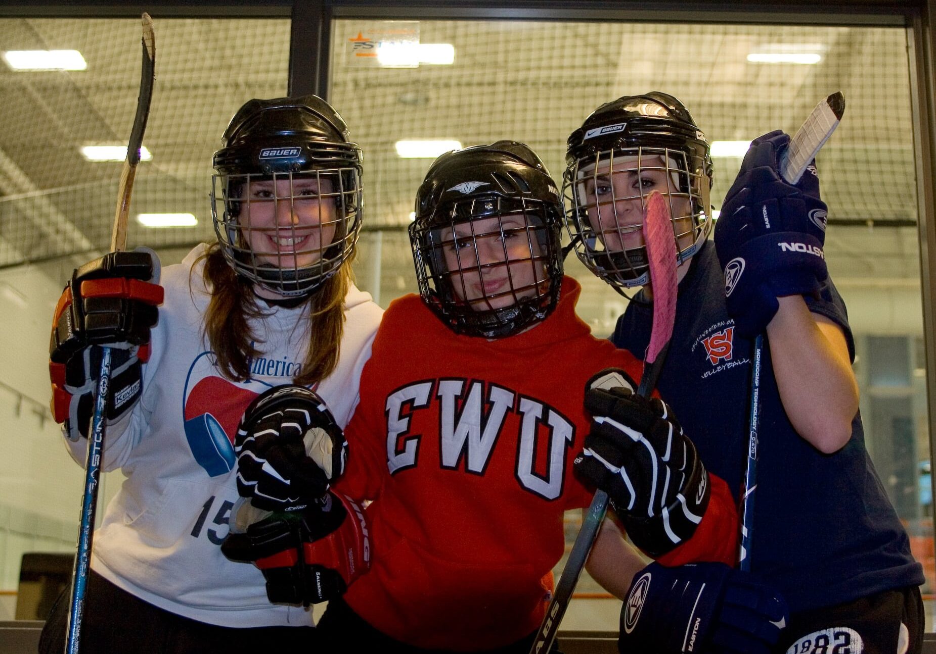 Baitlyn, Kayla, and Daitlin pose, wearing hockey gear, in front of the rec center's ice rink