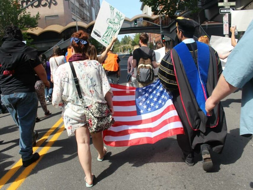 people taking place in a march. Two people are holding an American flag. The photo is from the back and is in Spokane