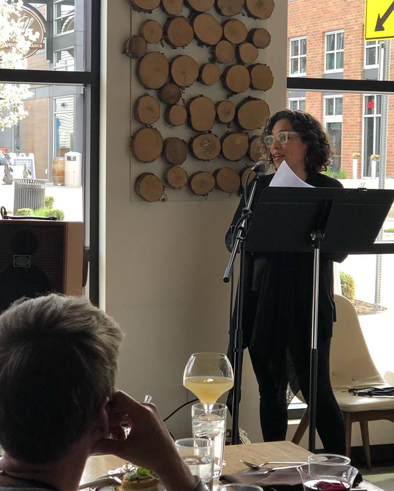 A photo of a woman standing with a metal stand reading poetry to a dining audience.