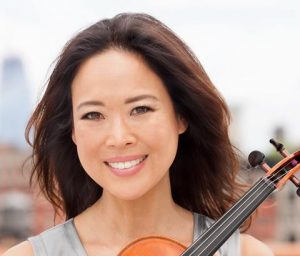 A person with brown hair and a violin smiles in an outdoor setting.