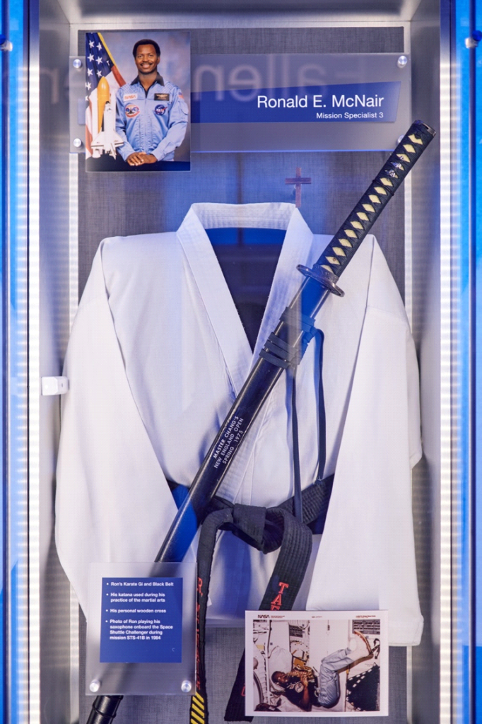 Museum exhibit from Kennedy Space Center memorial to those lost in te Challenger. A martial arts uniform is shown behind glass along with a sword, as well as two pictures of Ronald E. McNair. One of him in his NASA uniform, the other of him playing the saxaphone in space.