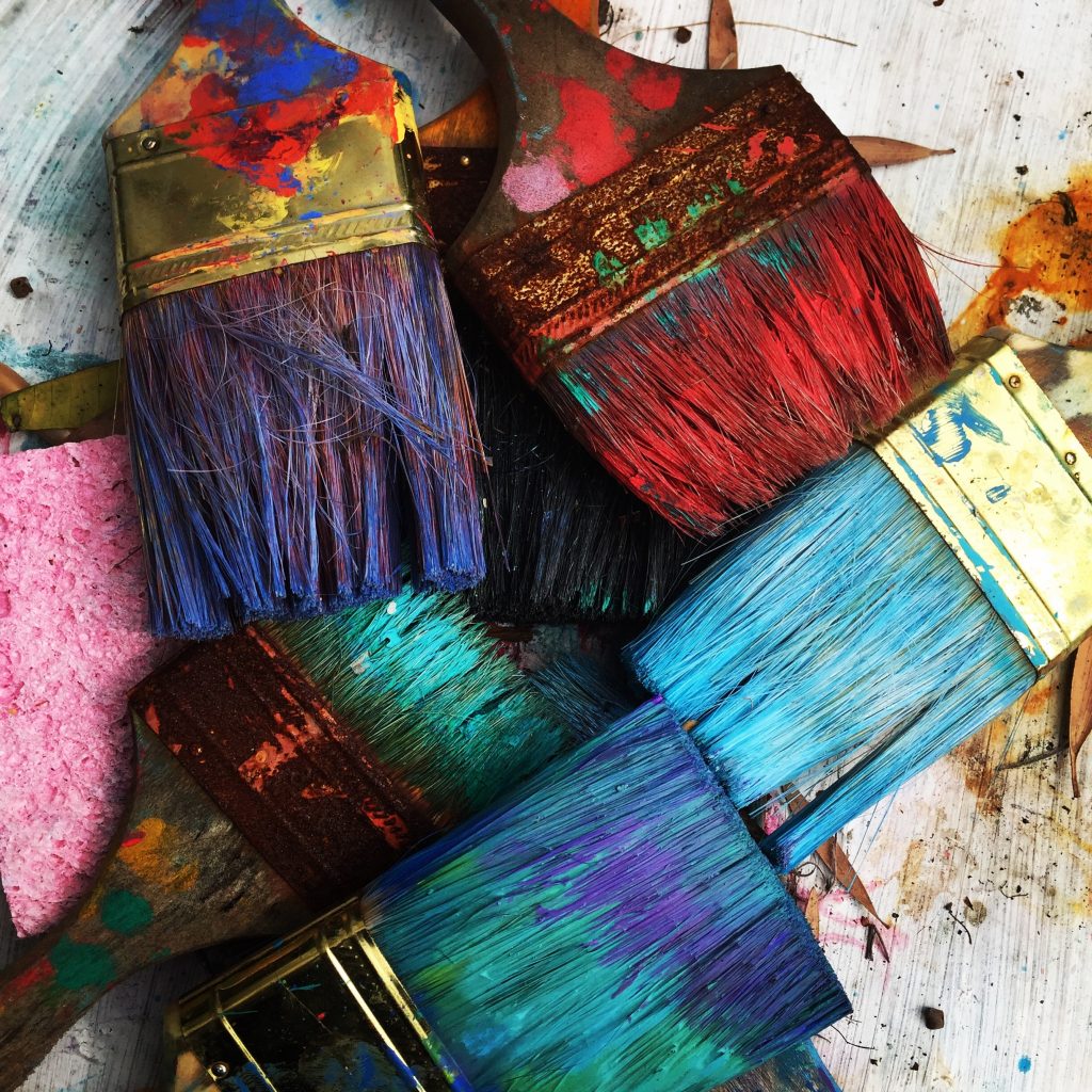 top down photo of paint brushes. They are covered in different colors and are laid on top of each other.