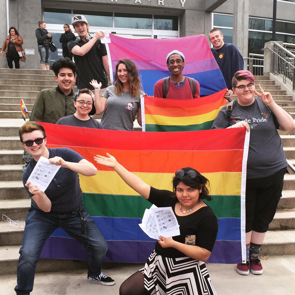 Students smiling on the JFK Library steps holding pride and bisexuality flags