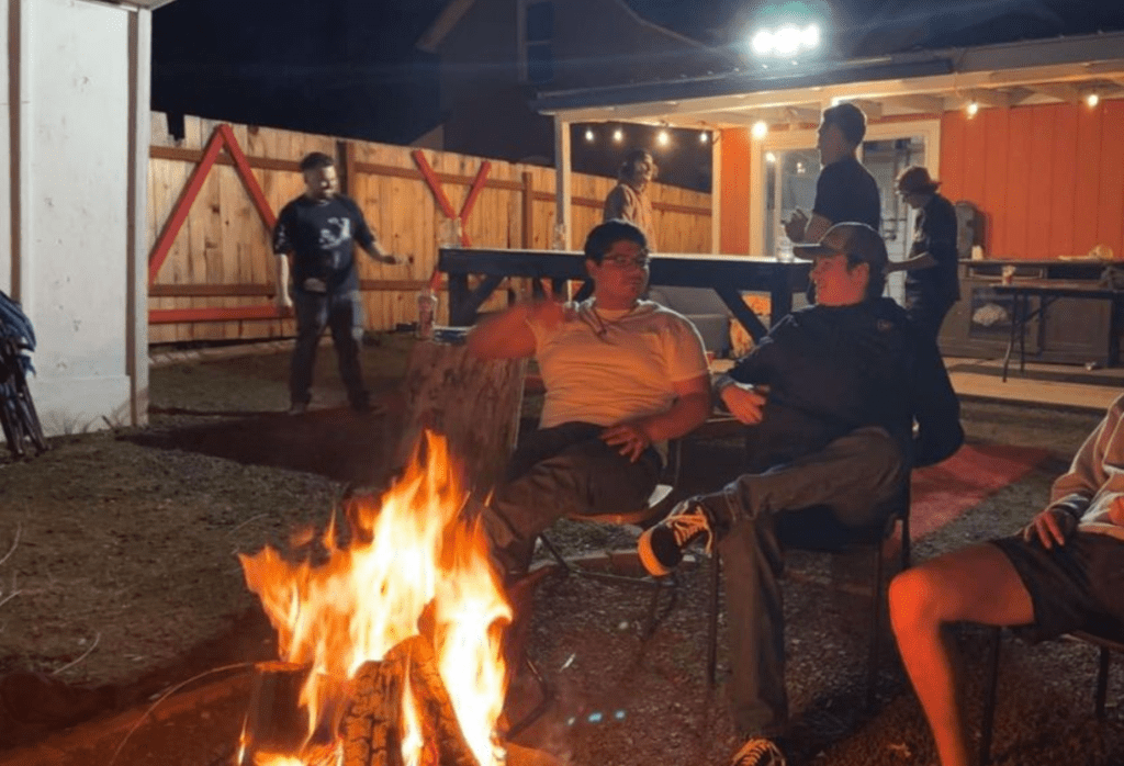 Students sitting around a campfire at night. Three students are in chairs while others are standing