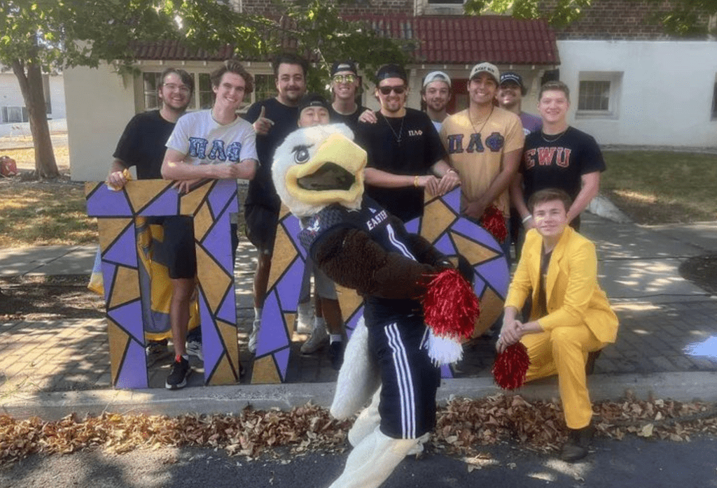 Swoop posing in front of fraternity house with fraternity members behind him