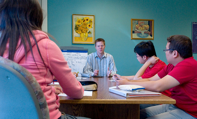 Man talks to group of students. They are sitting at a table within a room
